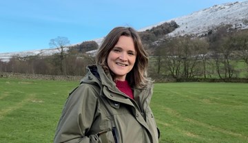Headshot of woman wearing outdoor clothing stood in a green field with a snow-covered hill behind.